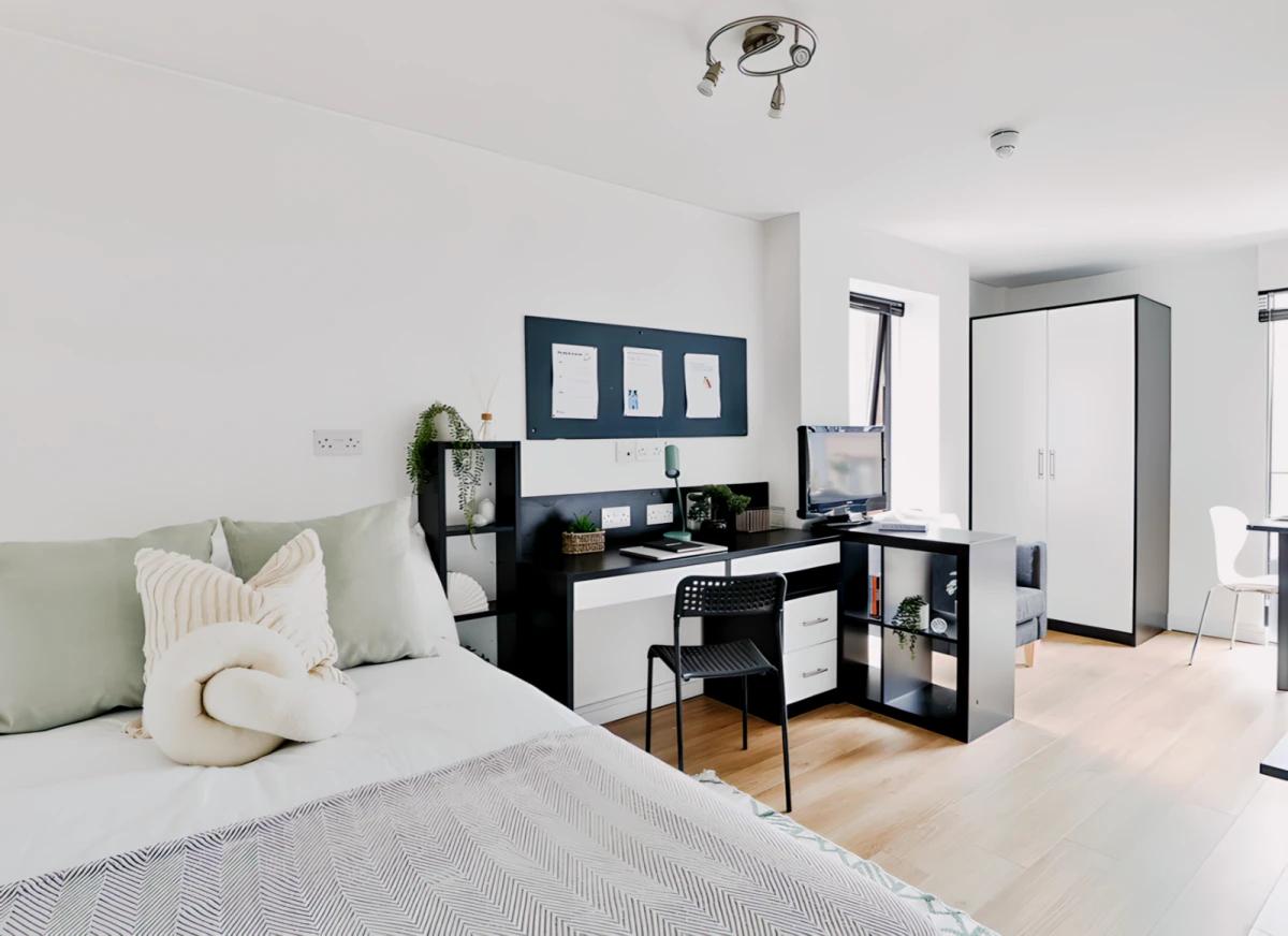 Bright modern studio room with a double bed, study desk, wardrobe, shelving, and contemporary furnishings.