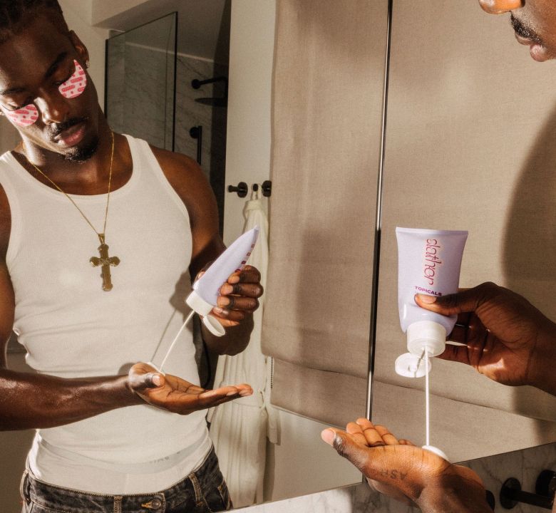 Man wearing pink eye patches pours product from a purple tube into his hand, reflected in a mirror.