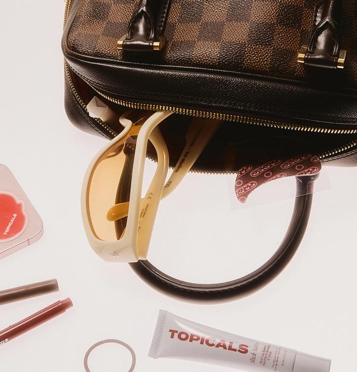 A Louis Vuitton handbag with cream sunglasses, a phone, makeup, and Topicals brand products on a white surface.
