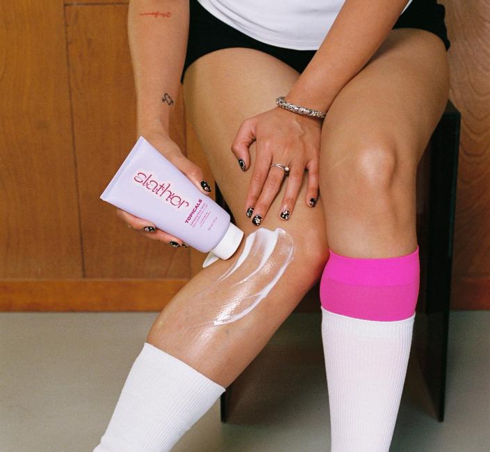 A person applies "Slather Topicals" lotion to their leg, wearing black shorts, white socks with a pink accent, and blue and pink heeled sandals.