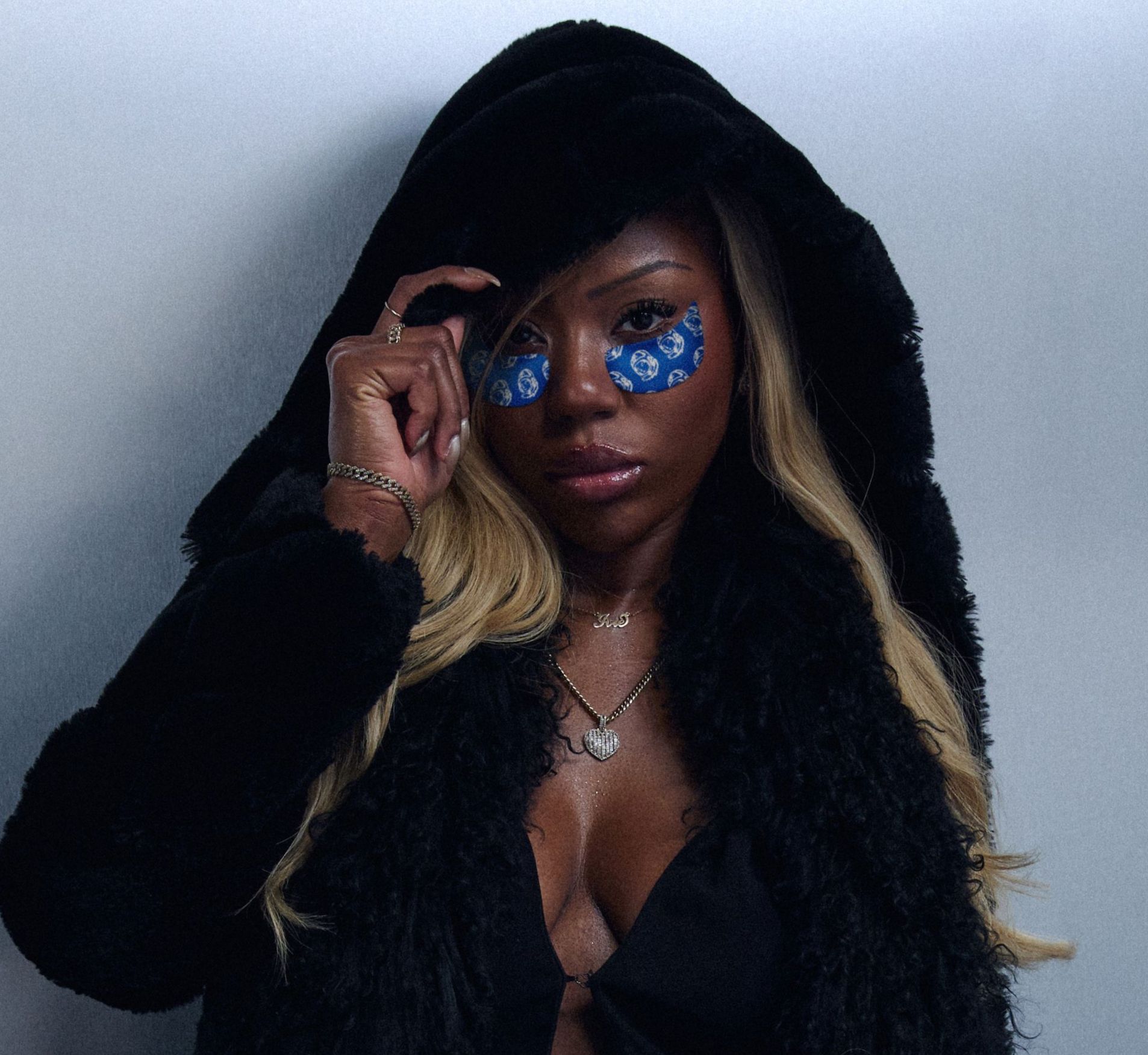 A woman in a black hooded coat with blonde hair wears blue patterned under-eye patches, looking directly at the viewer.