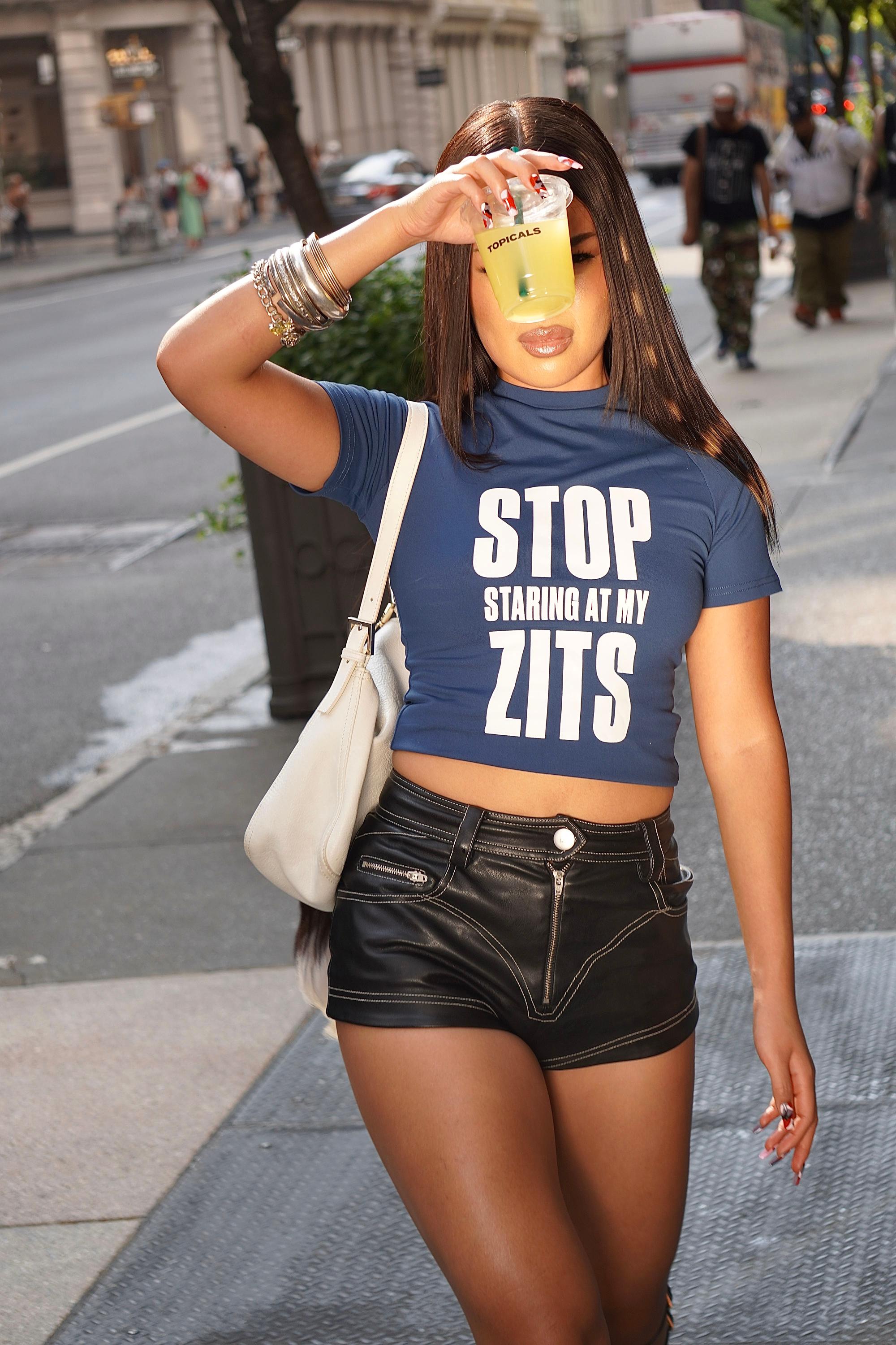 A woman in a blue crop top with "STOP STARING AT MY ZITS" printed on it and black shorts stands on a city sidewalk, holding a yellow drink in front of her face.