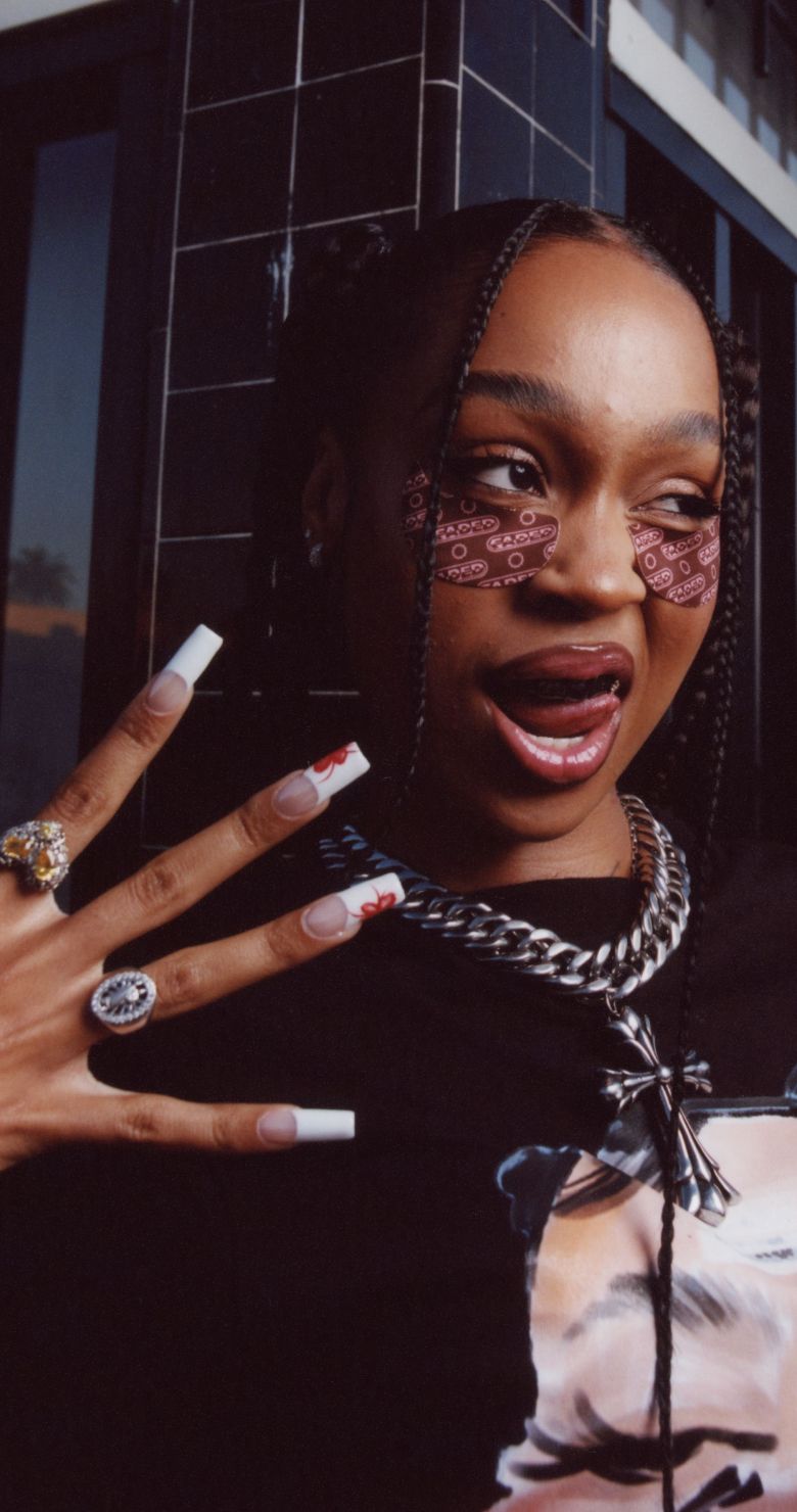 A young woman with braided hair and patterned under-eye patches makes a playful face, showing off long decorated nails and jewelry.