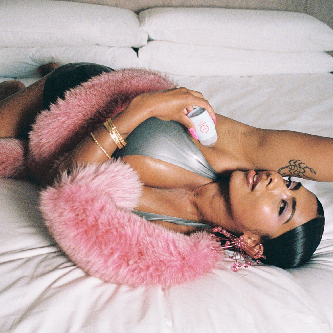 A woman lying on a white bed applies deodorant to her armpit, wearing a silver top and a pink fur boa.