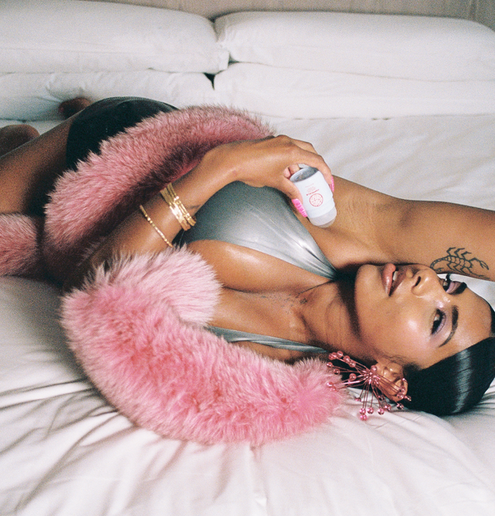 A woman lying on a white bed applies deodorant to her armpit, wearing a silver top and a pink fur boa.
