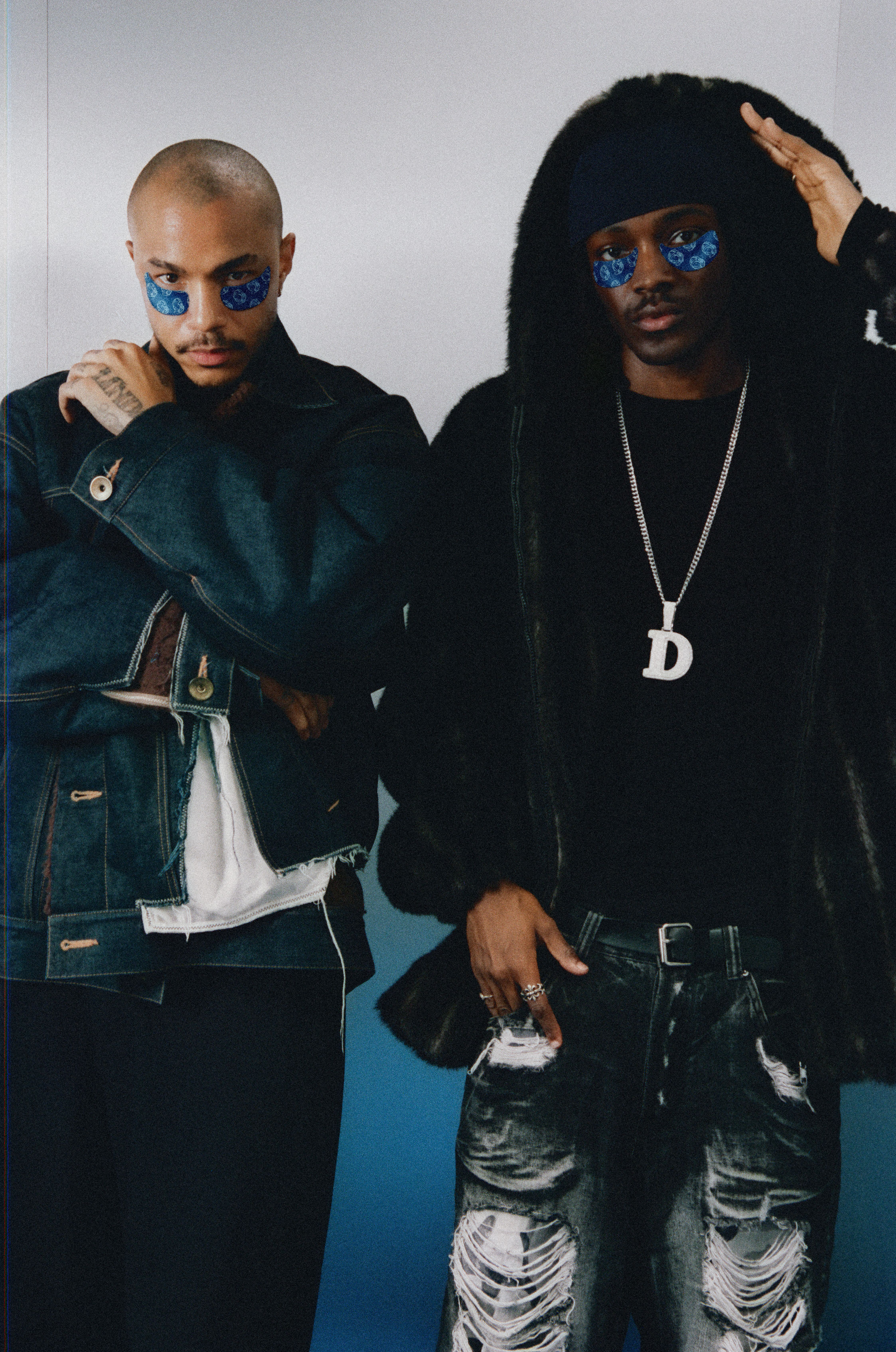 Two men with blue under-eye patches. The left man wears a denim jacket; the right man wears a dark fur-trimmed hooded jacket, ripped jeans, and a 'D' pendant.