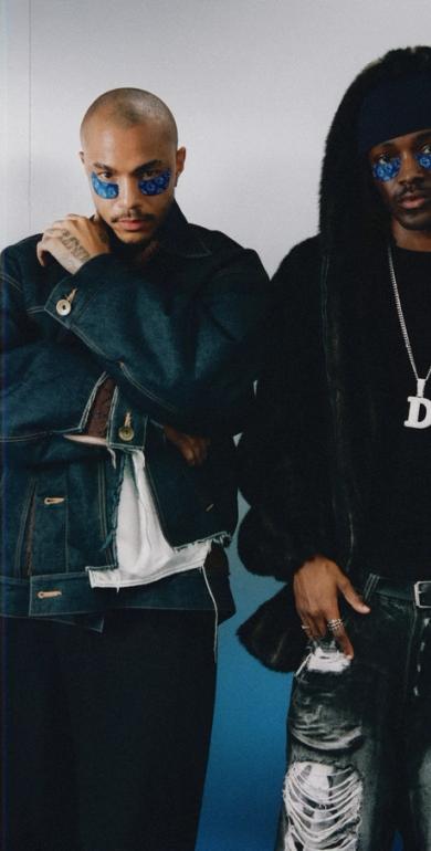 Two men with blue patterned eye adornments. One wears a denim jacket, the other a fur-trimmed jacket, ripped jeans, and a "D" pendant.