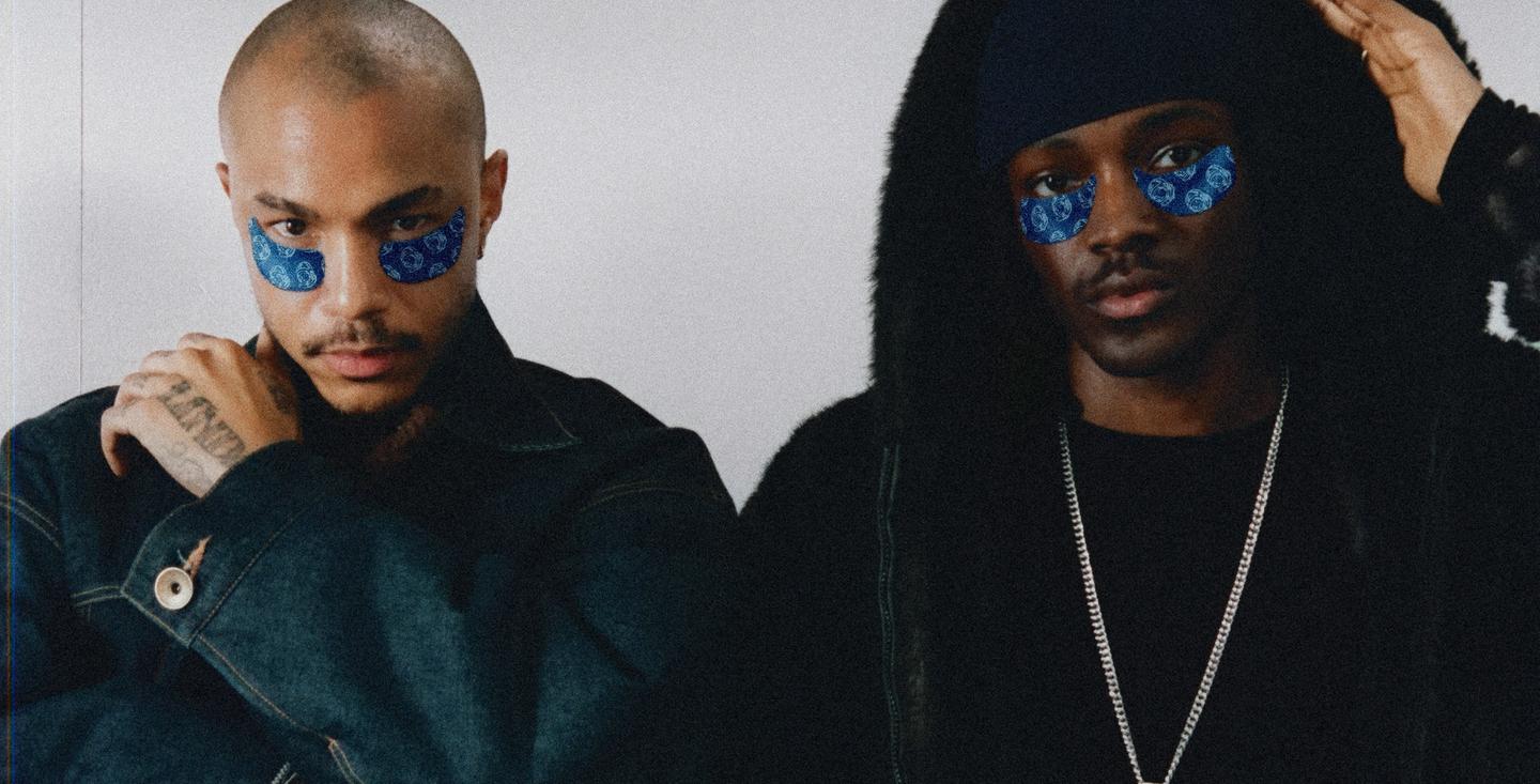 Two men with blue under-eye patches. The left man wears a denim jacket; the right man wears a dark fur-trimmed hooded jacket, ripped jeans, and a 'D' pendant.