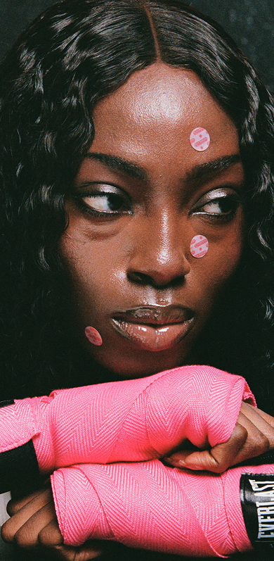 A person in a camouflage hooded jacket with Stüssy eye patches holds a hand out towards the viewer.