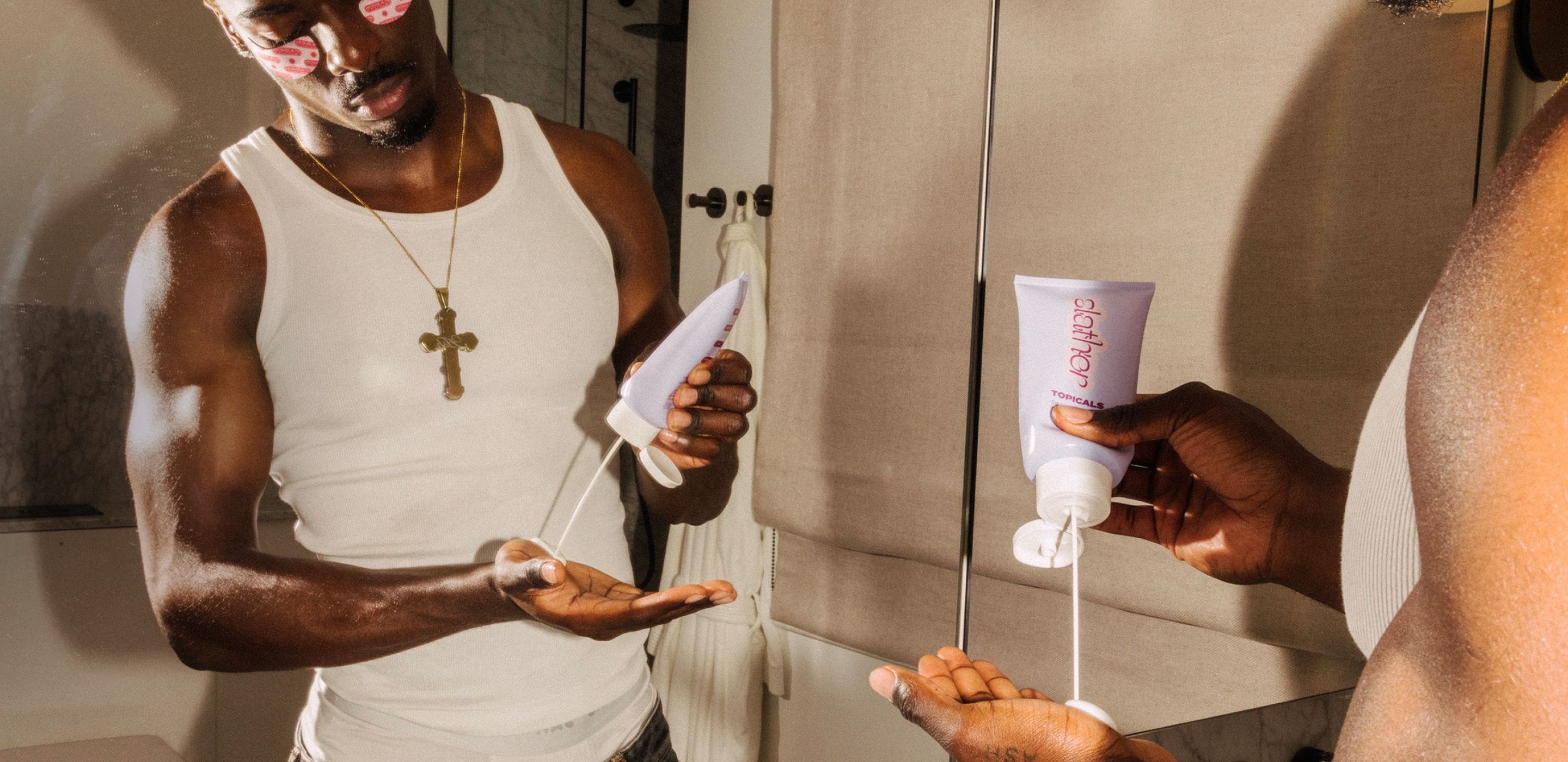 Man with eye patches pours lotion from a tube into his hand, reflected in a mirror.