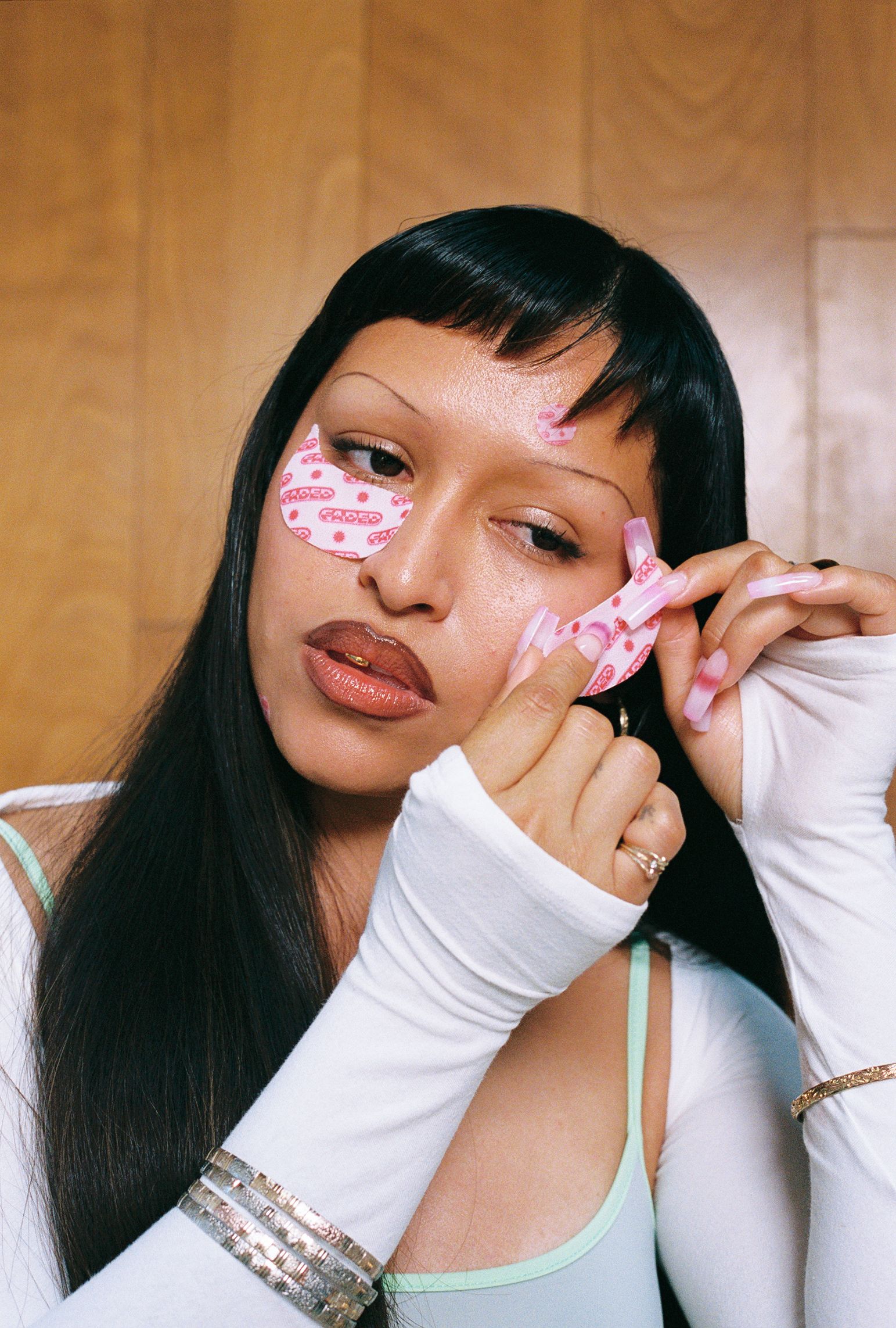 A person with long black hair and short bangs applies a pink patterned eye mask, displaying long pink acrylic nails, thin eyebrows, glossy lips with a gold tooth gem, and silver bracelets.