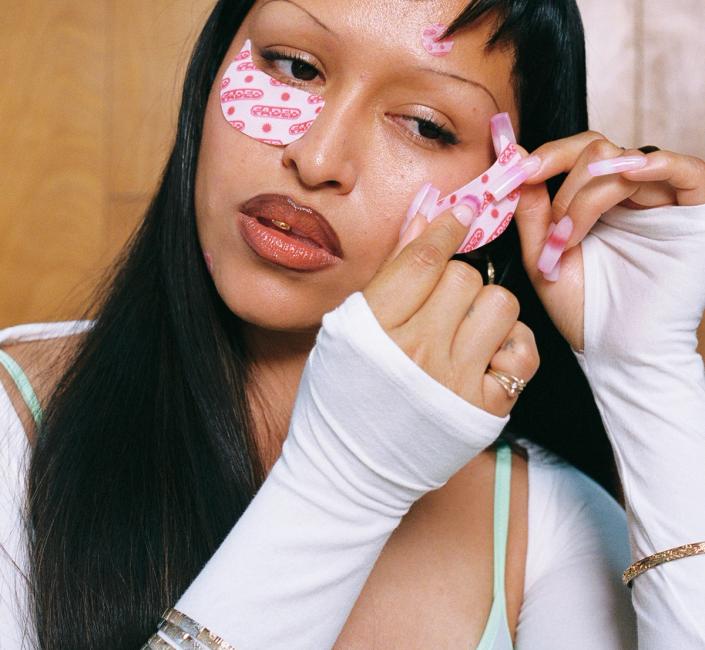A person with long black hair and short bangs applies a pink patterned eye mask, displaying long pink acrylic nails, thin eyebrows, glossy lips with a gold tooth gem, and silver bracelets.