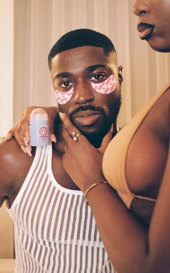 A man with a beard and pink eye patches looks forward, while a woman's hands hold a blue skincare product near him and touch his face.