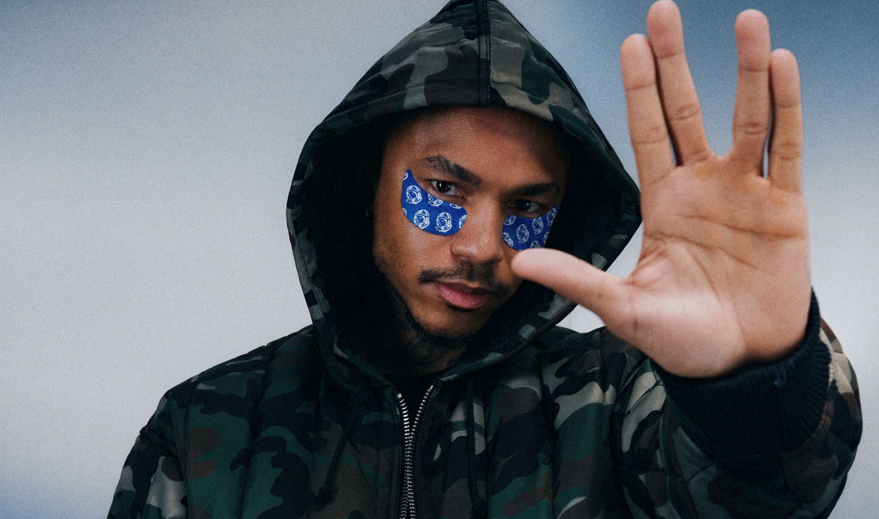 Man in camo hoodie and blue patterned eye patches performs a Vulcan salute.