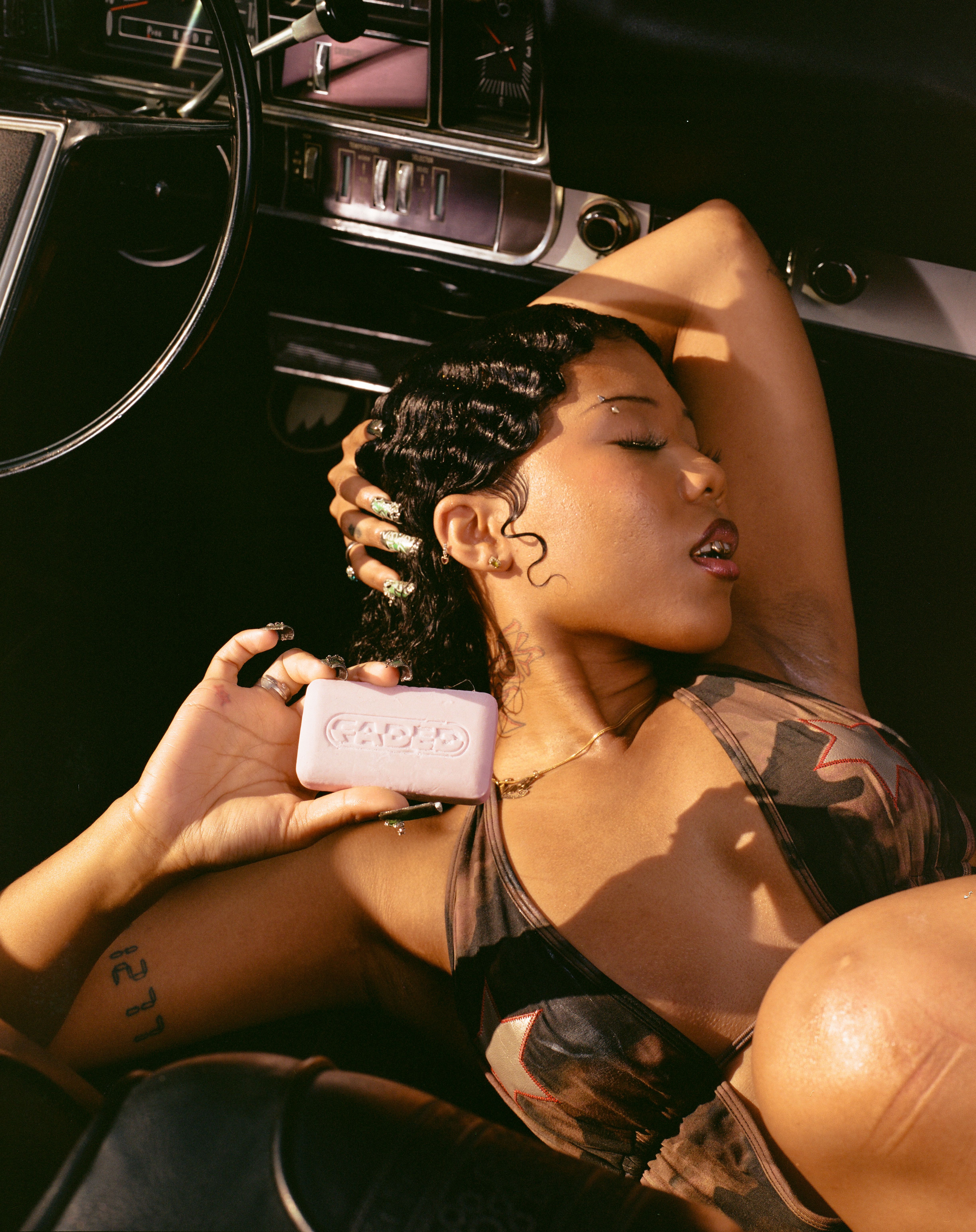 A woman with finger waves reclines in a vintage car, holding a pink bar of Cadde soap with her eyes closed.