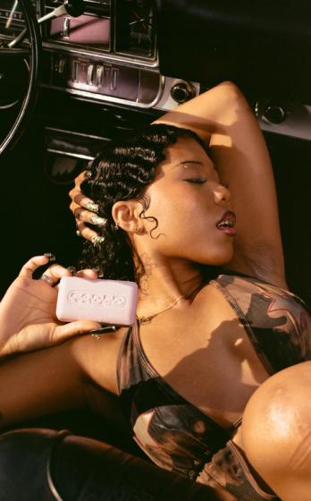 A woman with finger waves reclines in a vintage car, holding a pink bar of Cadde soap with her eyes closed.