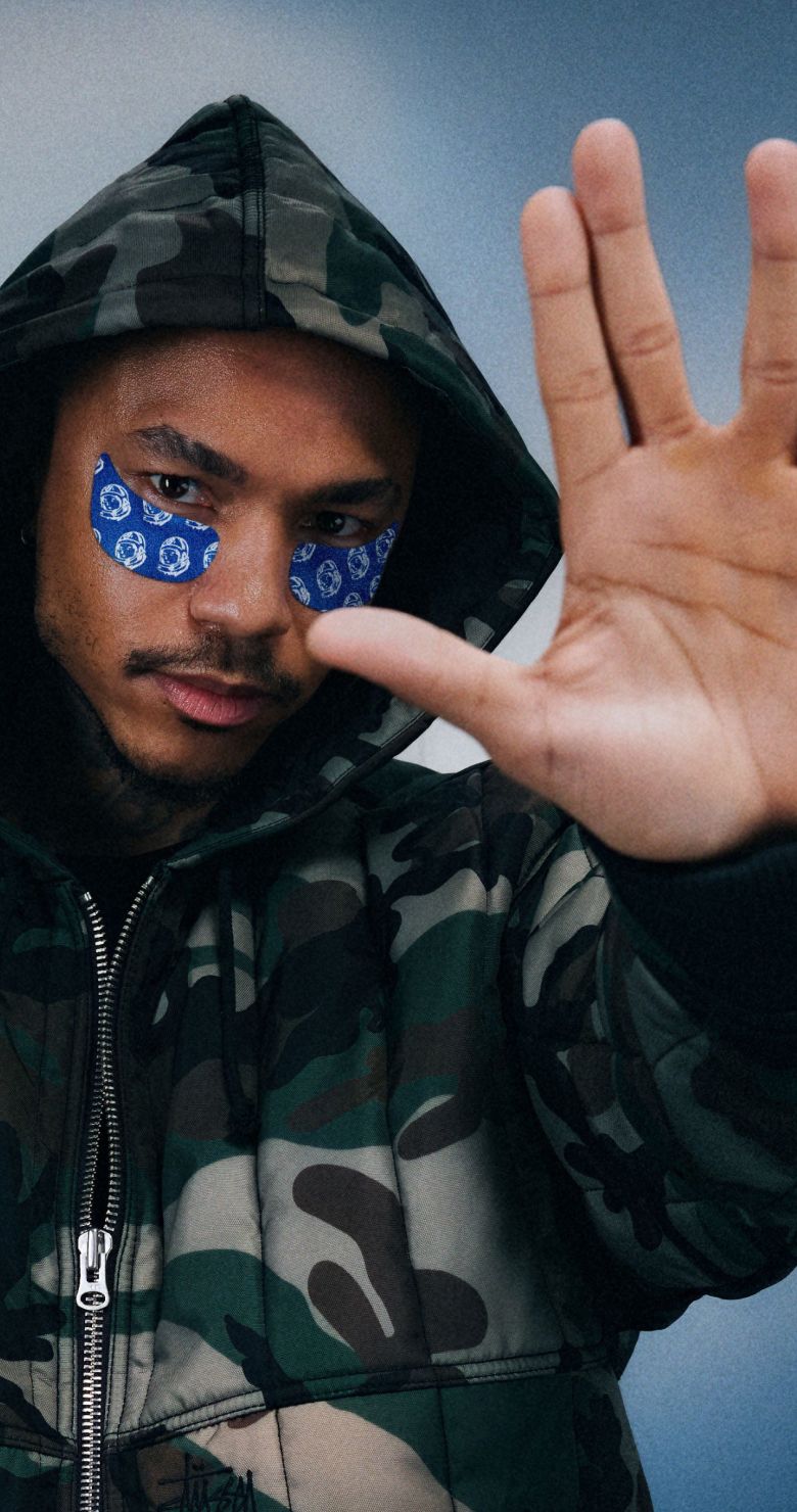A man in a camouflage hooded jacket with blue eye patches raises his palm towards the viewer.
