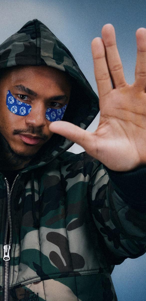 A man in a camouflage hooded jacket with blue eye patches raises his palm towards the viewer.