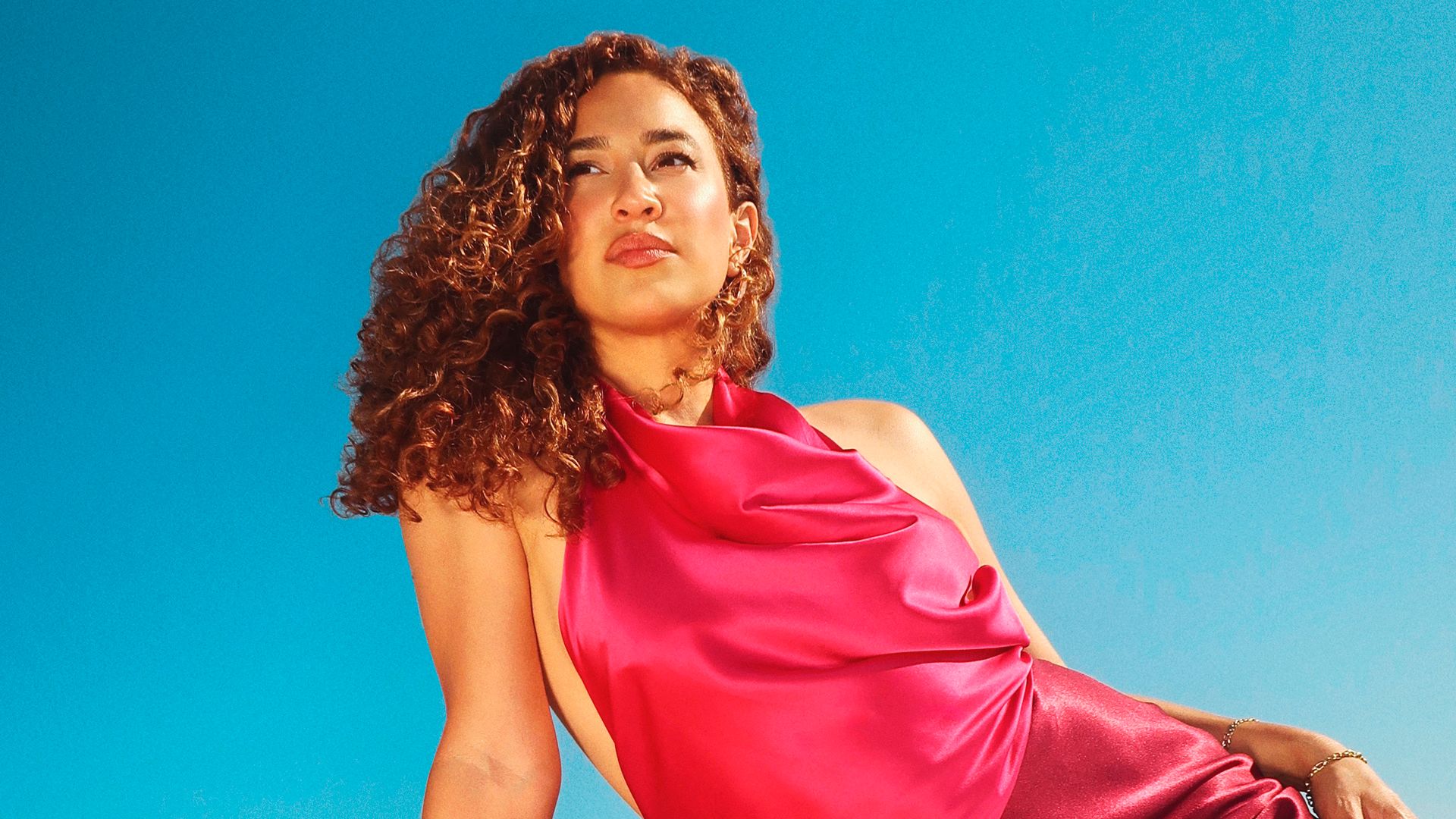 Woman with curly brown hair in a pink satin dress looking up against a bright blue sky.