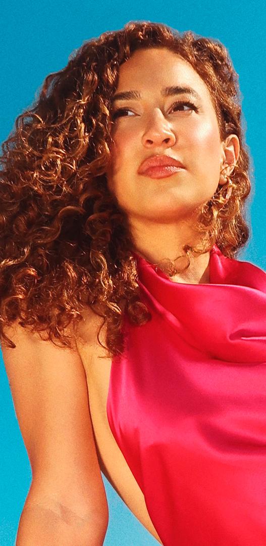 A woman with curly brown hair in a pink satin dress looks up against a blue sky.