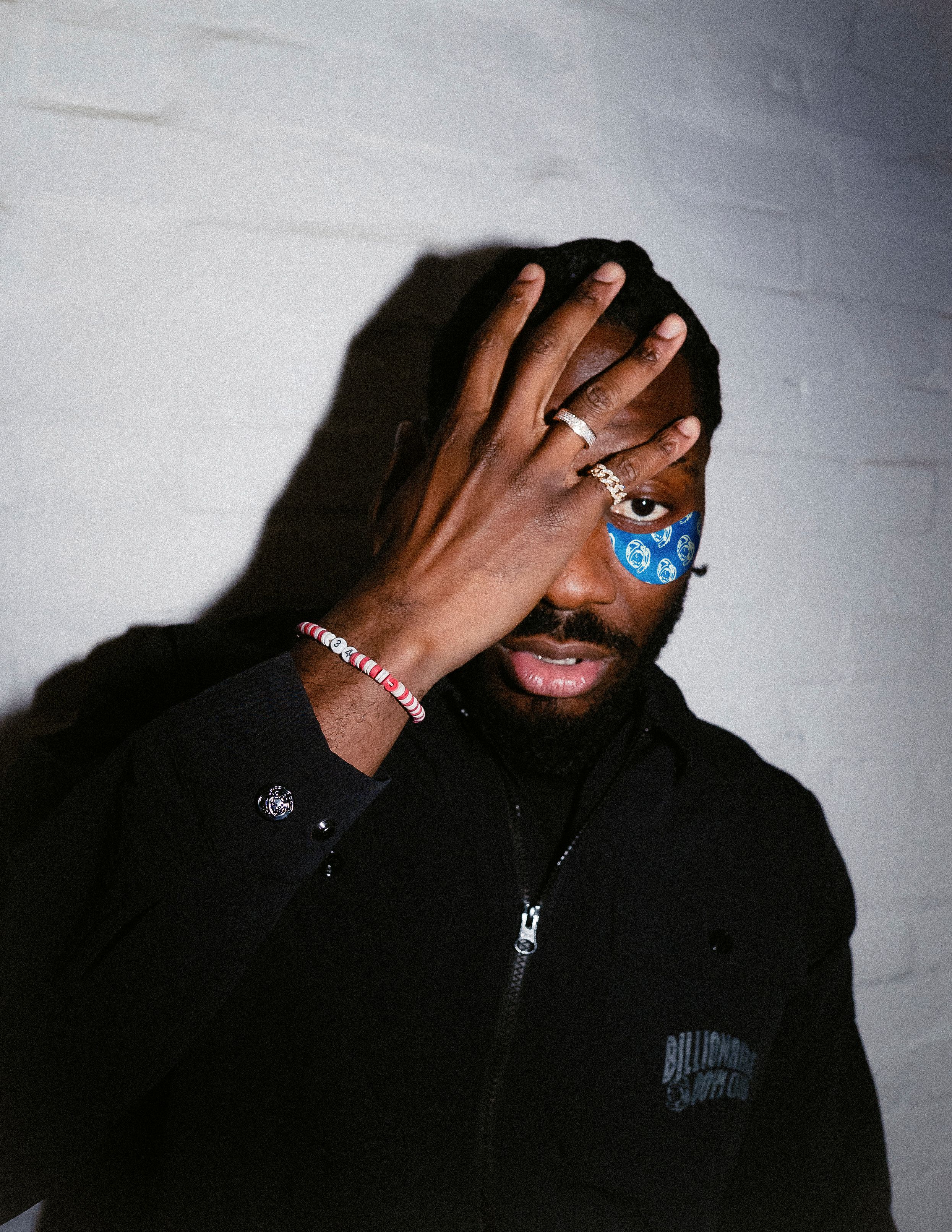 A man with his right hand covering his right eye, revealing a blue patterned under-eye patch below his left eye. He wears rings and a beaded bracelet, looking forward.