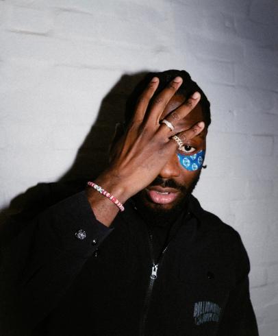 A man with his right hand covering his right eye, revealing a blue patterned under-eye patch below his left eye. He wears rings and a beaded bracelet, looking forward.