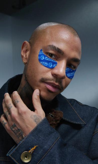 A man with a shaved head and tattoos wears a denim jacket and blue "GADED" eye patches, his hand resting on his chin, looking at the viewer.