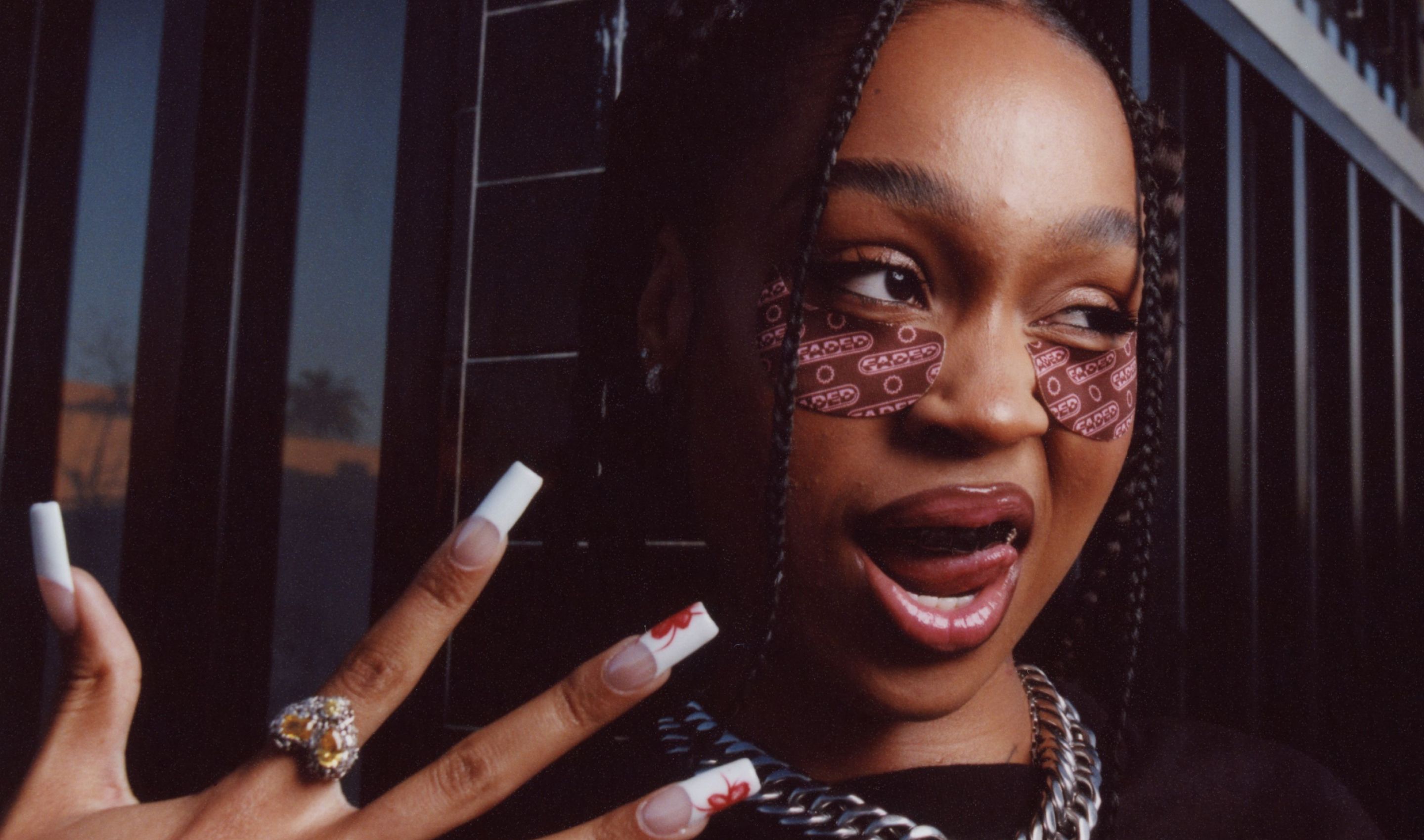 A young woman with braided hair, patterned eye patches, and long ornate nails playfully sticks out her tongue.