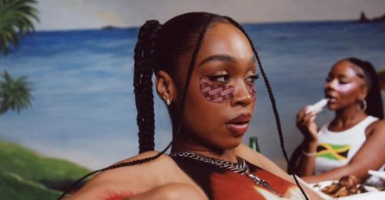 A Black woman with braids wears pink 'Kardashian' under-eye masks, with another person blurred against a tropical background.