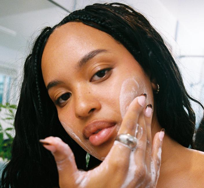 A woman with braids applies a foamy cleanser to her cheek, looking at the camera.