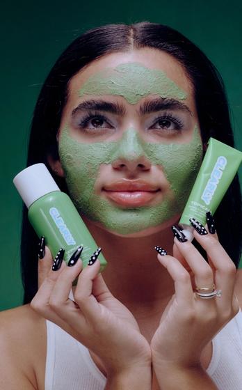 Young woman with a green face mask holds two green Clearly Topical skincare products against a green background.