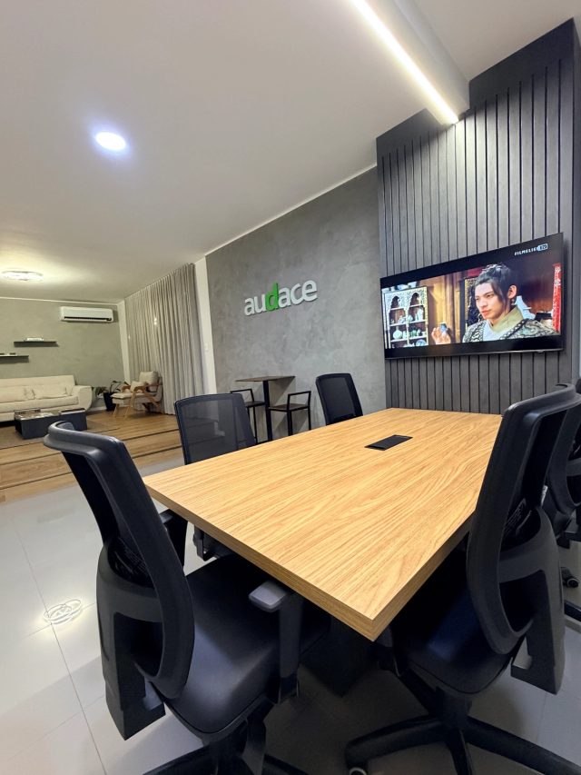 Sala de reunião da Audace: mesa de madeira com cadeiras pretas, televisão na parede ripada e o logo da agência na parede de cimento queimado