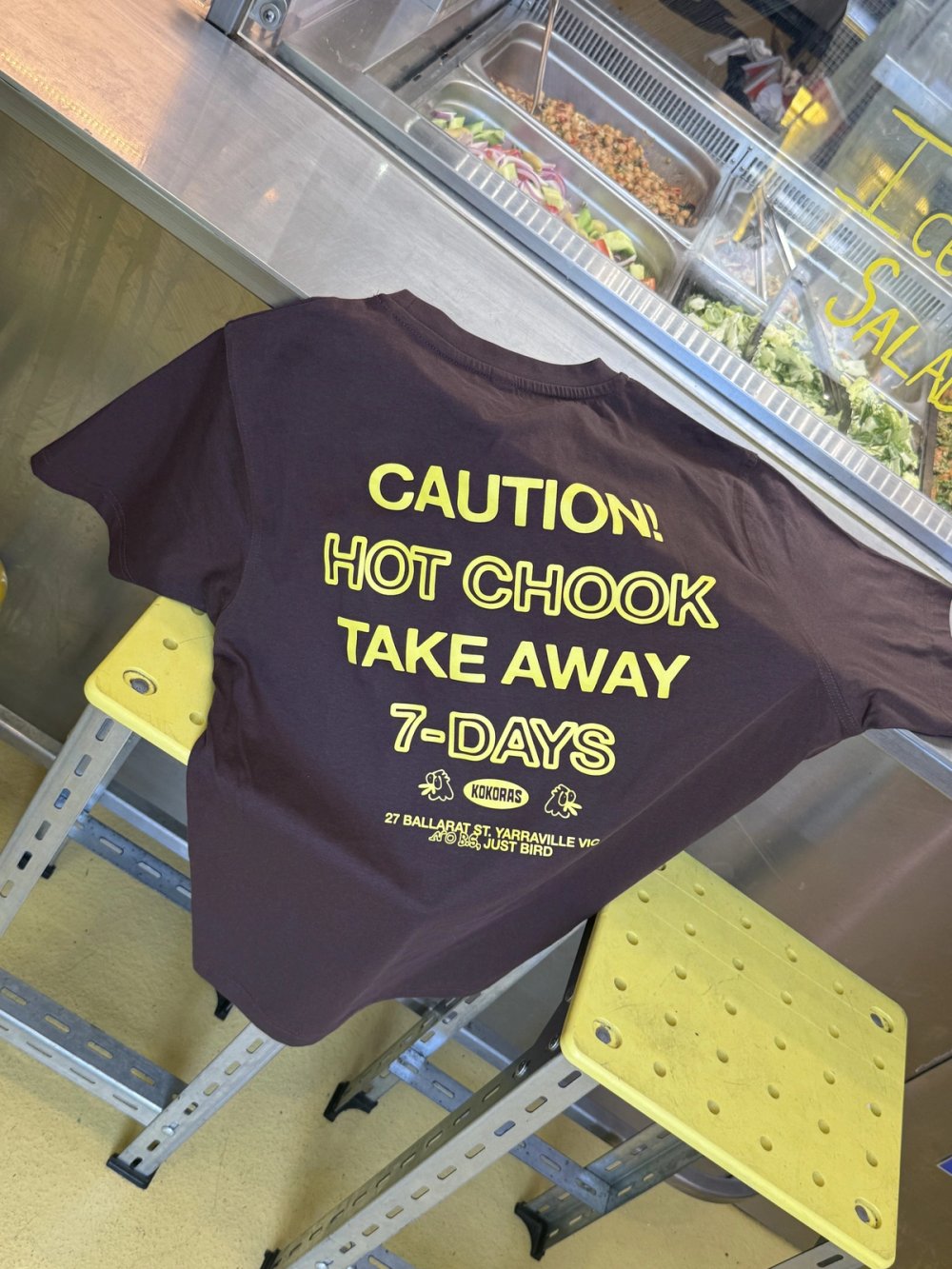 Kokoras "Caution! Hot chook take away" T-shirt draped over a yellow stool