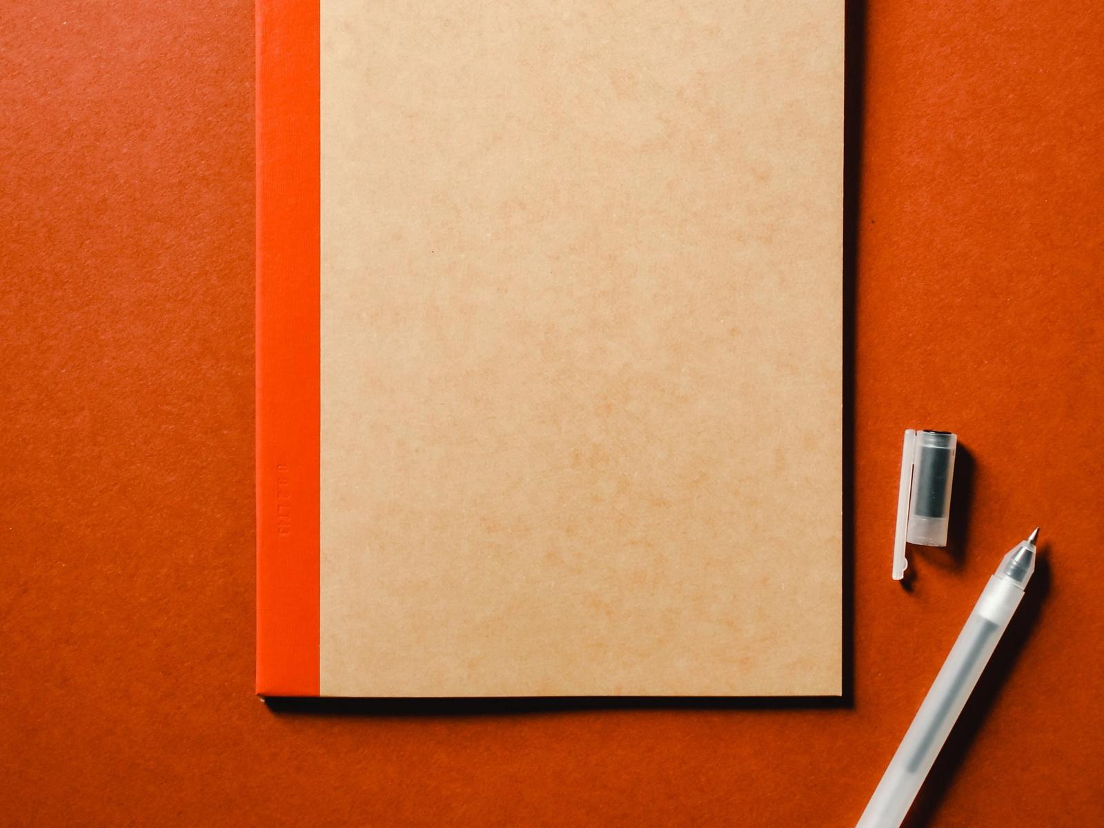 A closed notebook and pen on an orange surface.