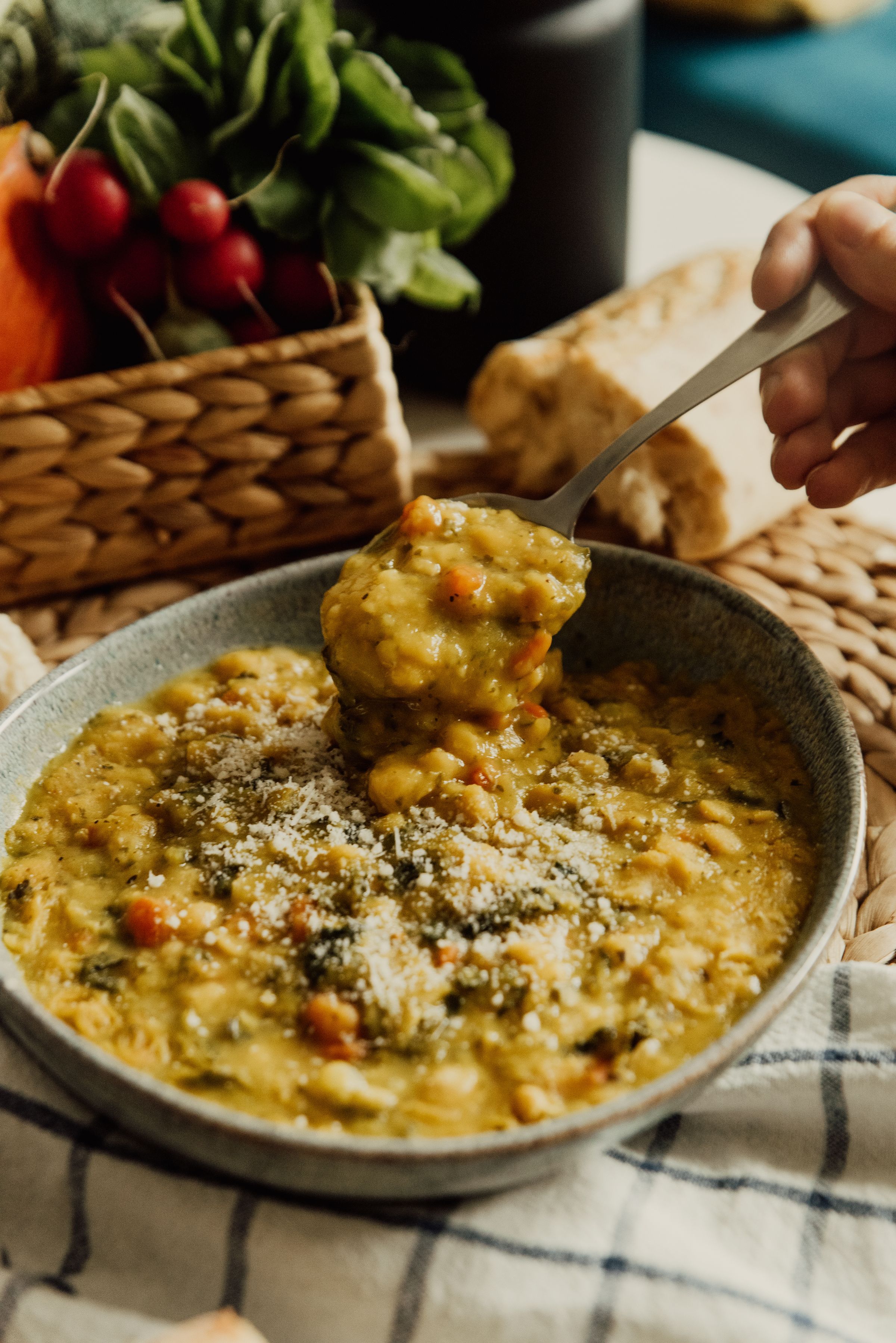 A spoon lifting thick soup from a bowl, with bread and vegetables nearby.