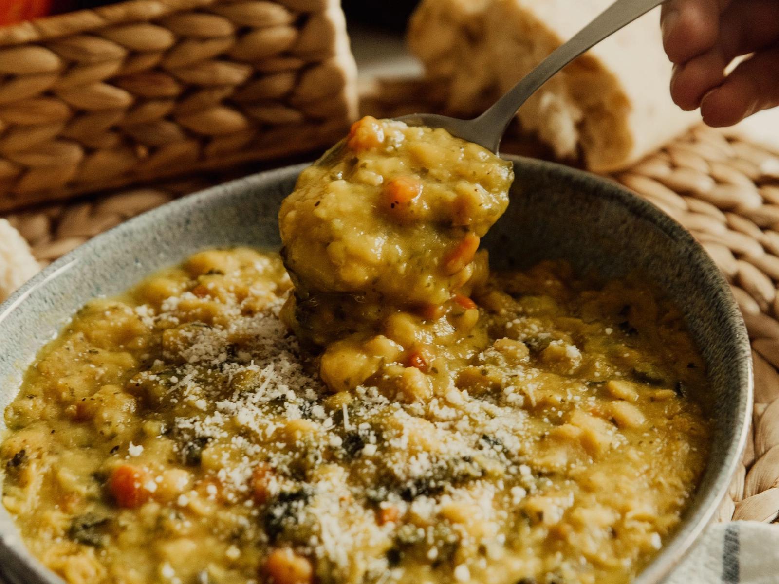 A spoon lifting thick soup from a bowl, with bread and vegetables nearby.