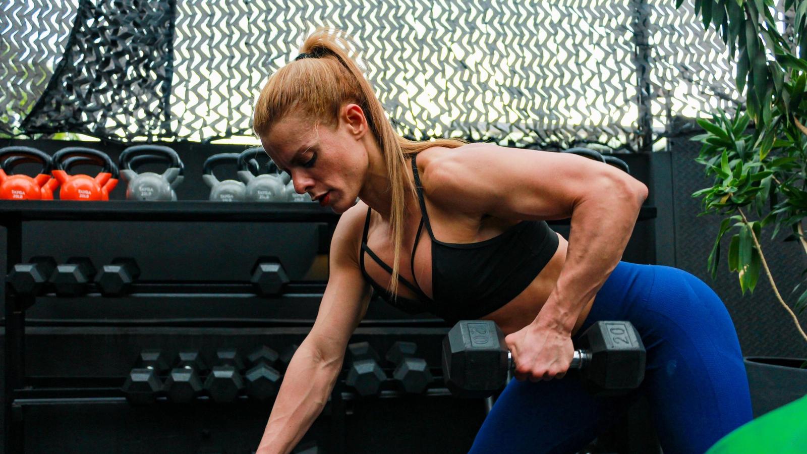 A woman performing a dumbbell row beside a gym bench.