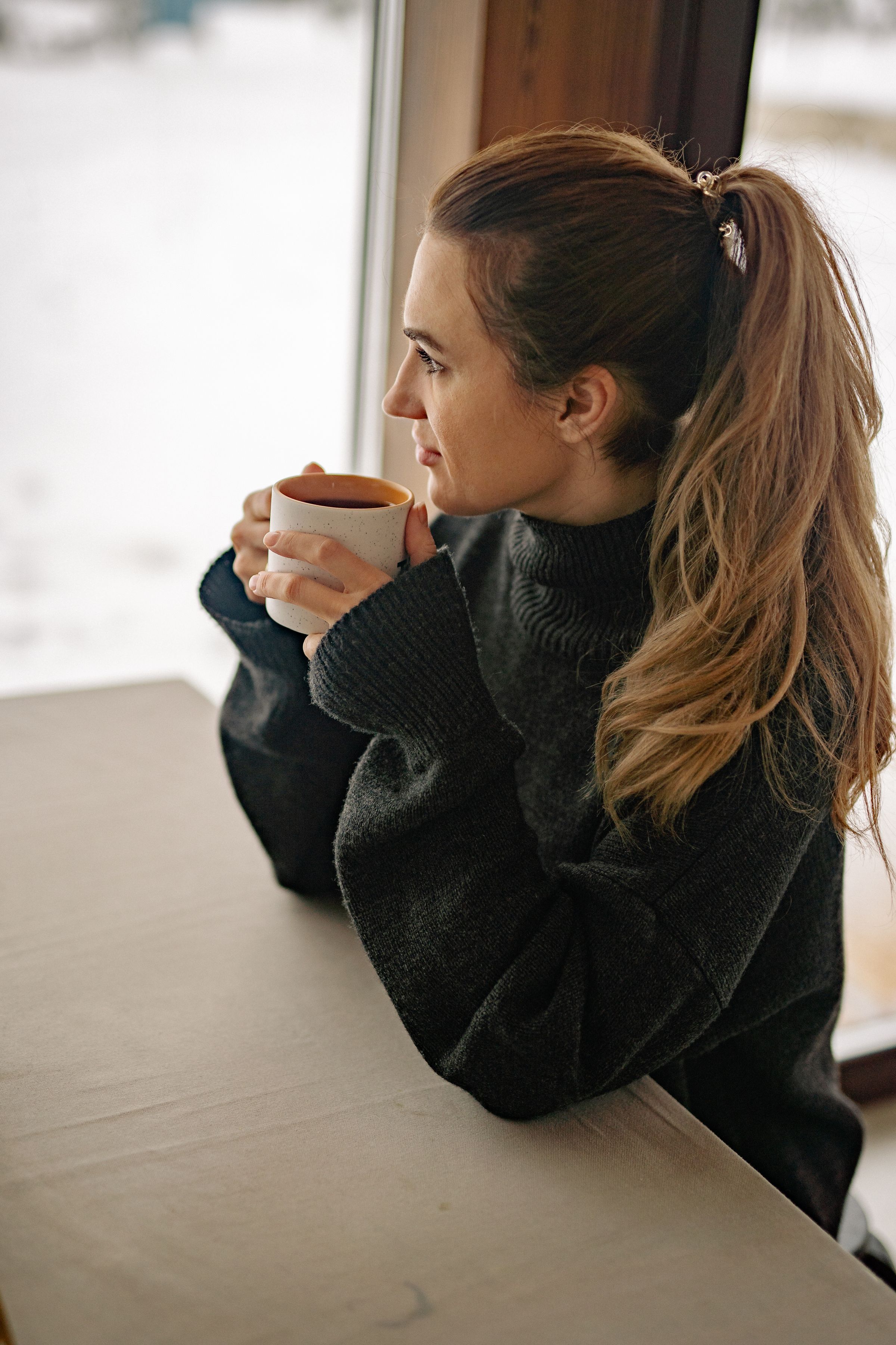A woman holding a mug beside a window.