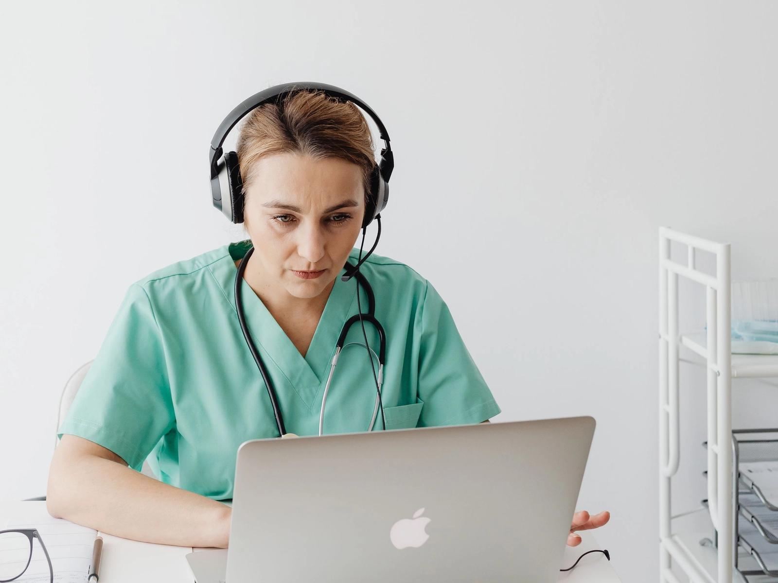 A woman in green scrubs using a laptop and headset.