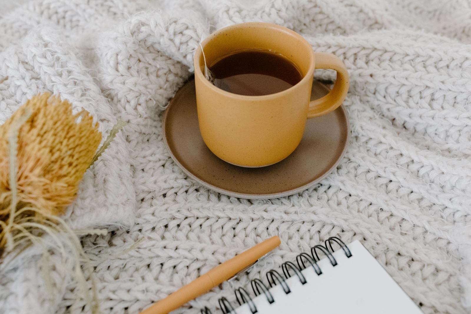 A yellow cup of tea beside a notebook on a knitted blanket.