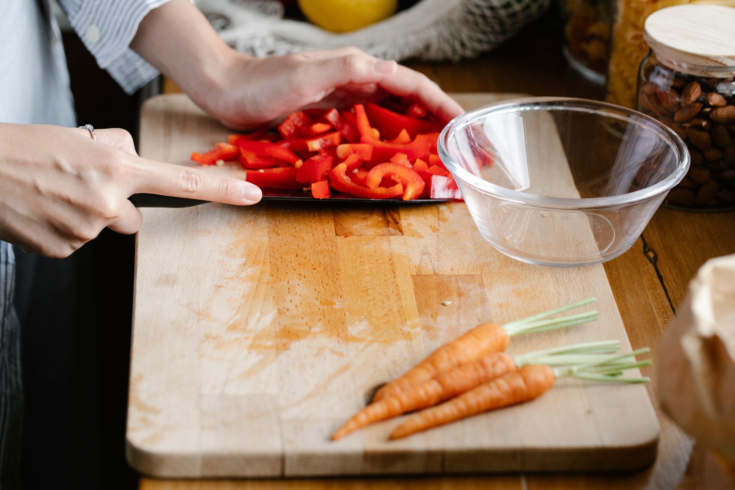 Hands cutting a red pepper on a board beside carrots.