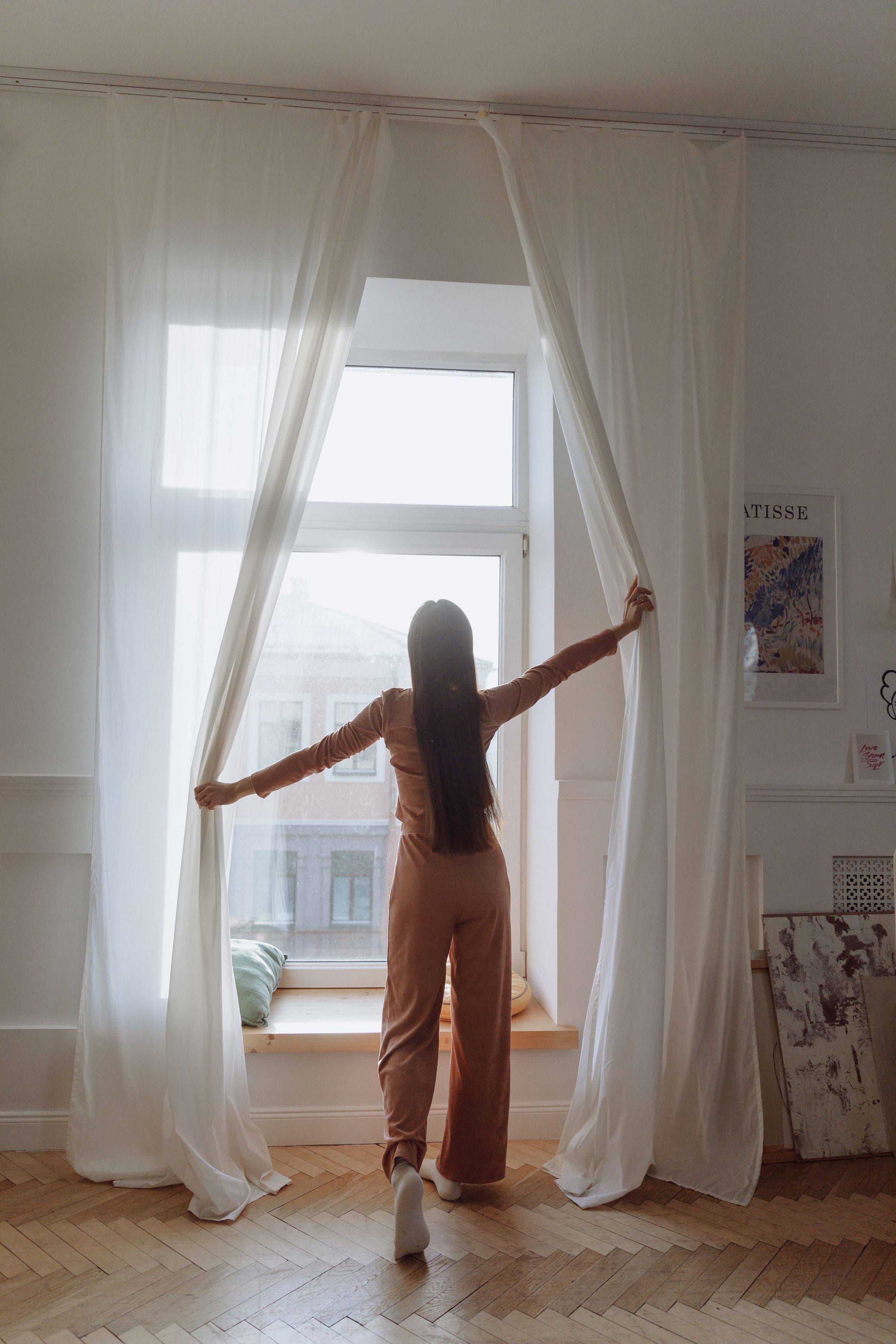 A woman opening curtains beside a bed.