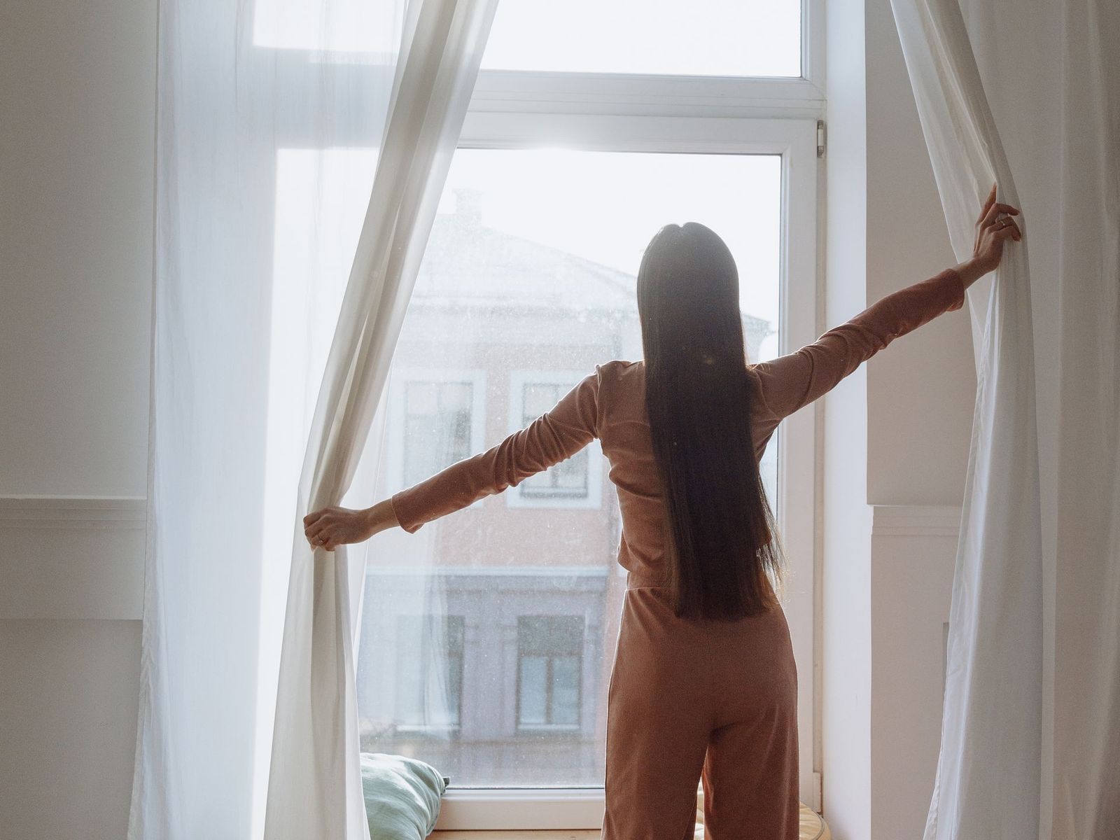 A woman opening curtains beside a bed.