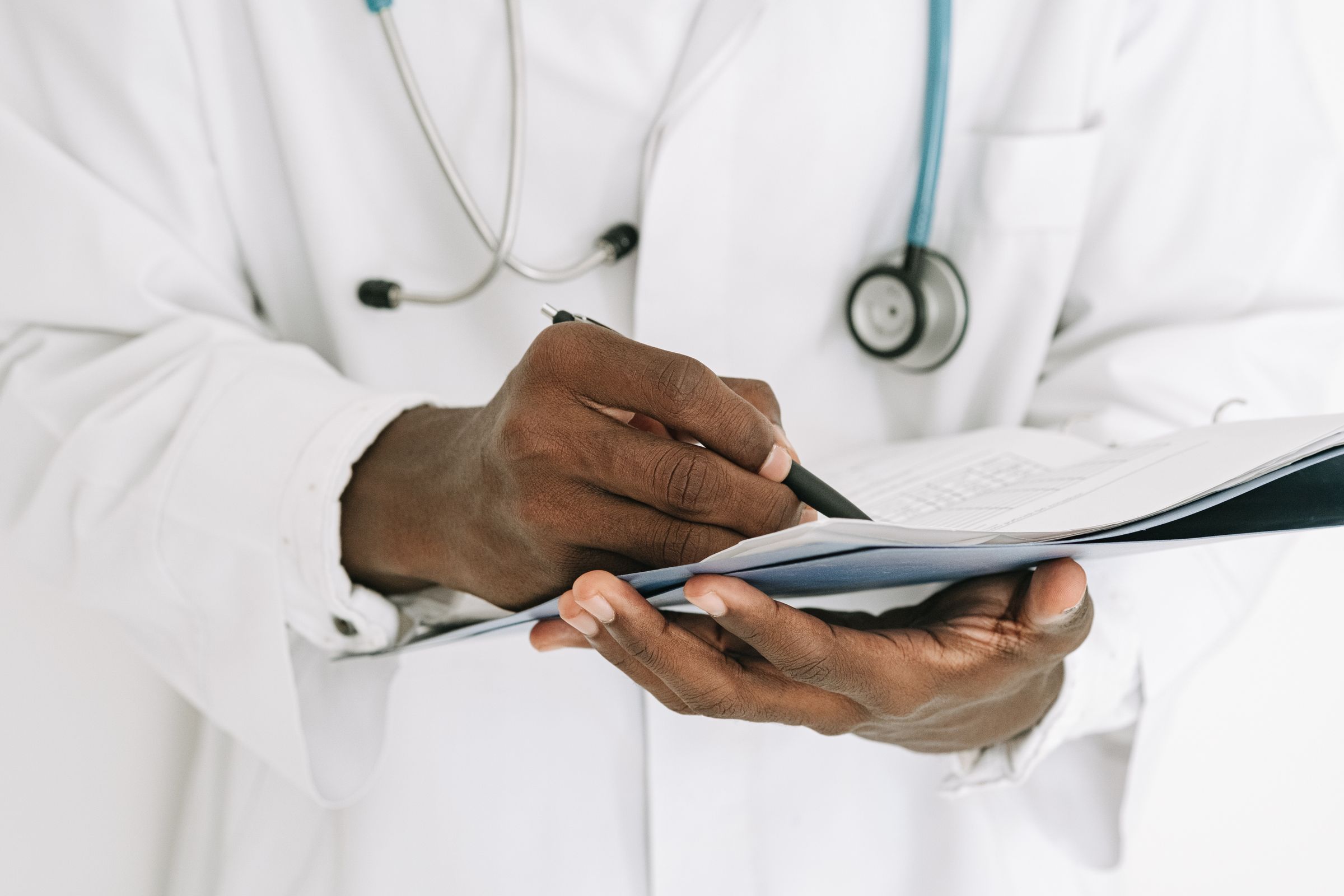 Hands writing on a clipboard, with a stethoscope over a white coat.