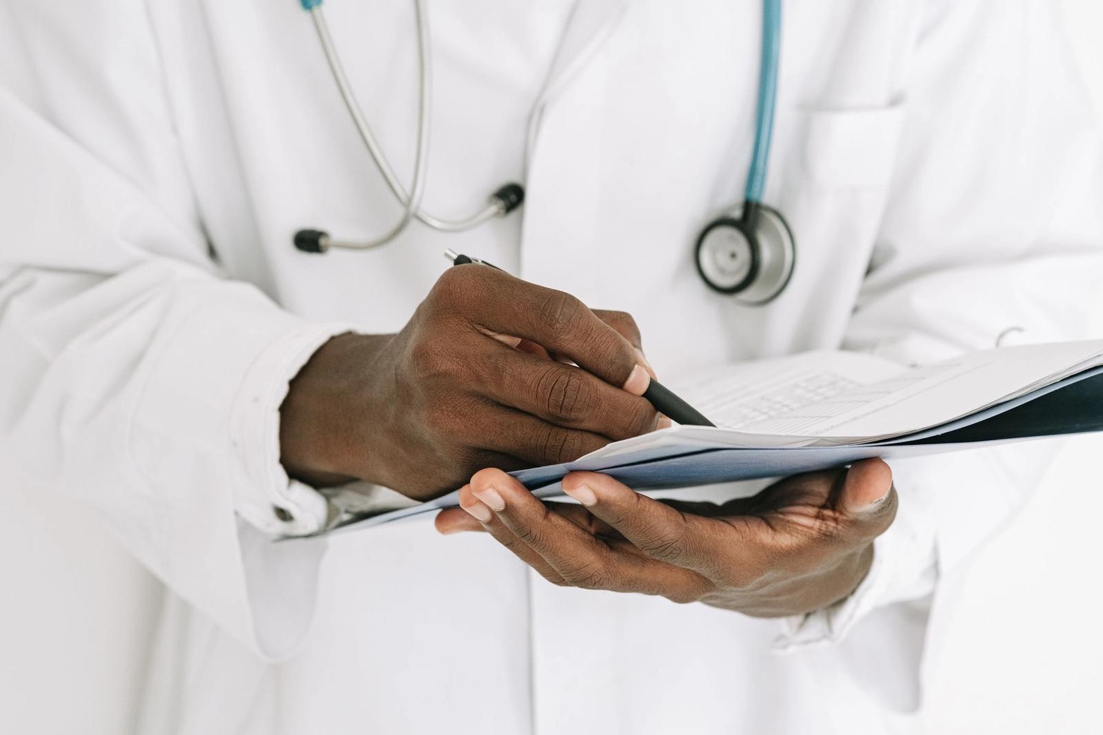 Hands writing on a clipboard, with a stethoscope over a white coat.