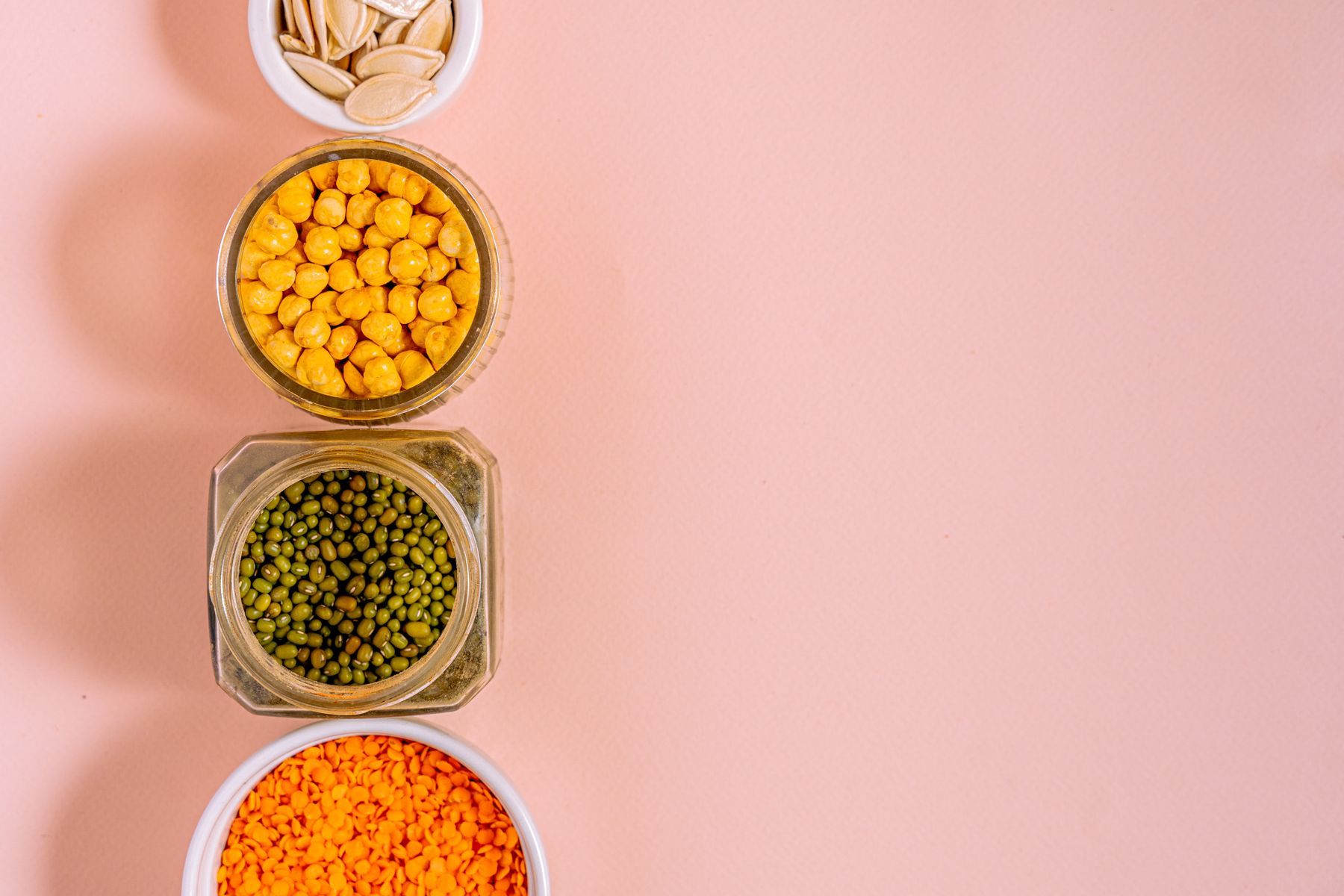 Bowls of chickpeas, lentils, beans and seeds on a pink surface.
