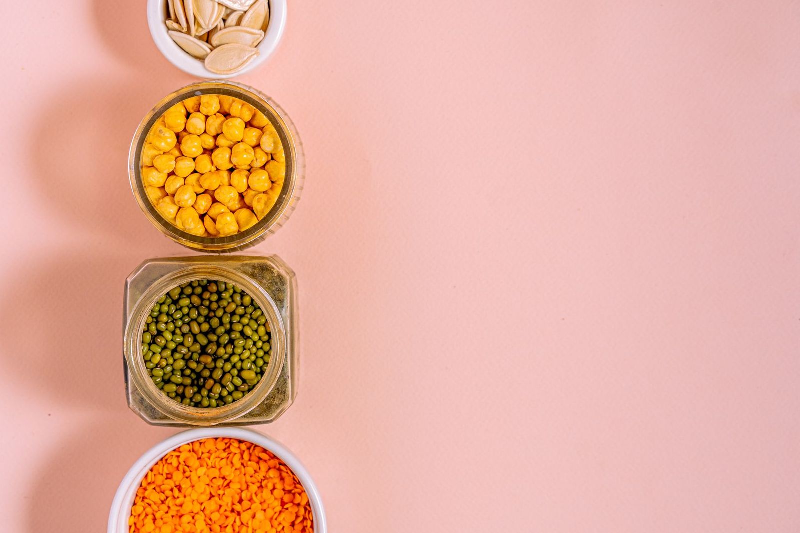 Bowls of chickpeas, lentils, beans and seeds on a pink surface.