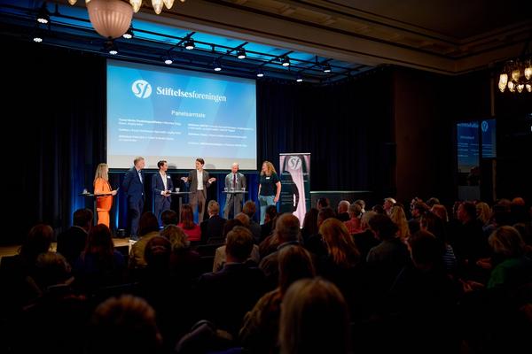 stor sal med mage mennesker, seks personer på scenen til panelsamtale