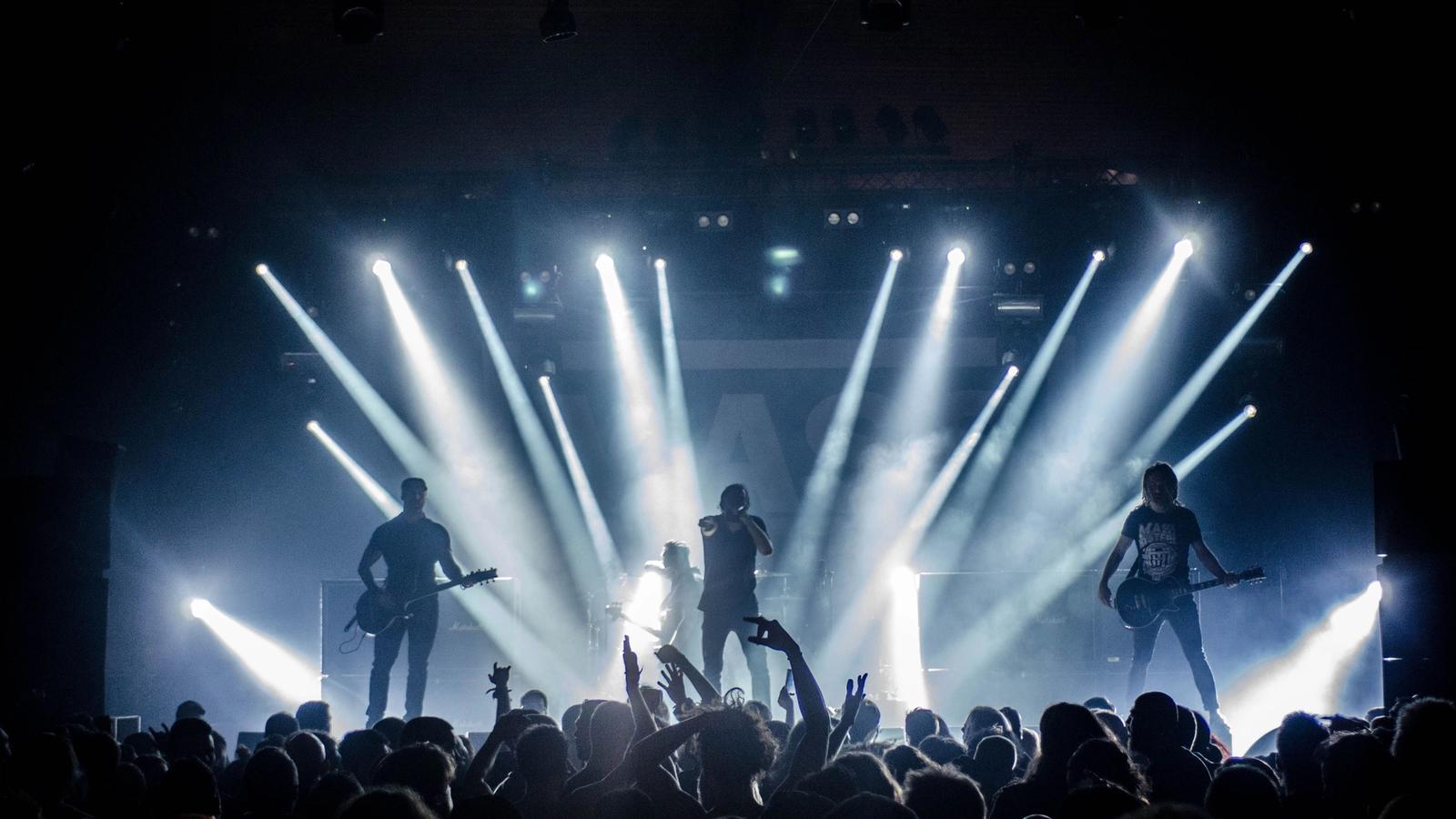 A four-piece band performs on a club stage, silhouetted against bright white light beams above a cheering crowd.