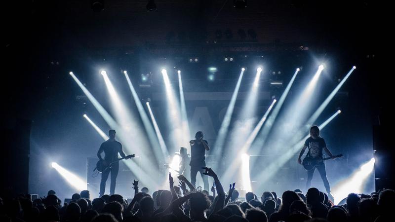 A four-piece band performs on a club stage, silhouetted against bright white light beams above a cheering crowd.